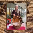 Smiling older woman holding woven baskets at an exhibit table on an outdoor patio. She also holds hand-woven cooking utensils. UC Davis Department of Plant Sciences