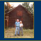 An older man and woman standing in front of an old-looking barn, smiling. The man has one arm around the woman.