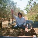 Woman stands on top of wagon in a field of almonds. She is holding a shovel and smiling at the camera.