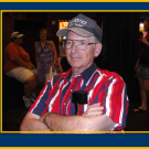 Older man in a ball cap and red-striped shirt, arms crossed, photographed at indoor social gathering
