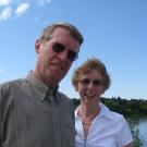 Older man and woman standing by a wide river. Sunny day, blue sky behind them. They are smiling.